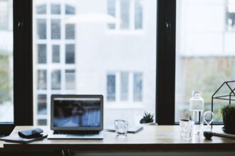 Desk setup by a large window with a laptop, notebook, glass, plant, and water bottle.