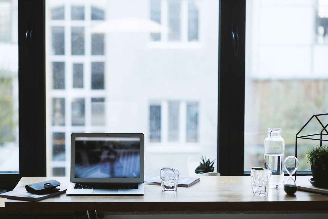 Desk setup by a large window with a laptop, notebook, glass, plant, and water bottle.
