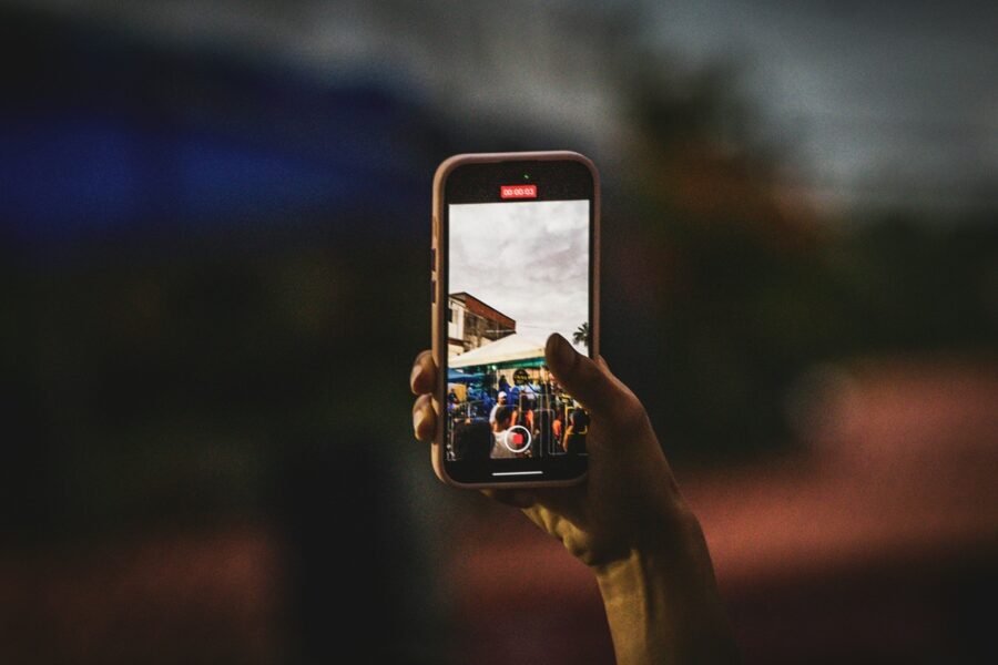Hand holding a smartphone, recording a crowded outdoor market or street fair seen on the screen.