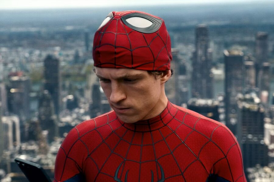 Spider-Man in a red web-pattern suit stands on a city rooftop, looking down at a phone against a blurred skyline.